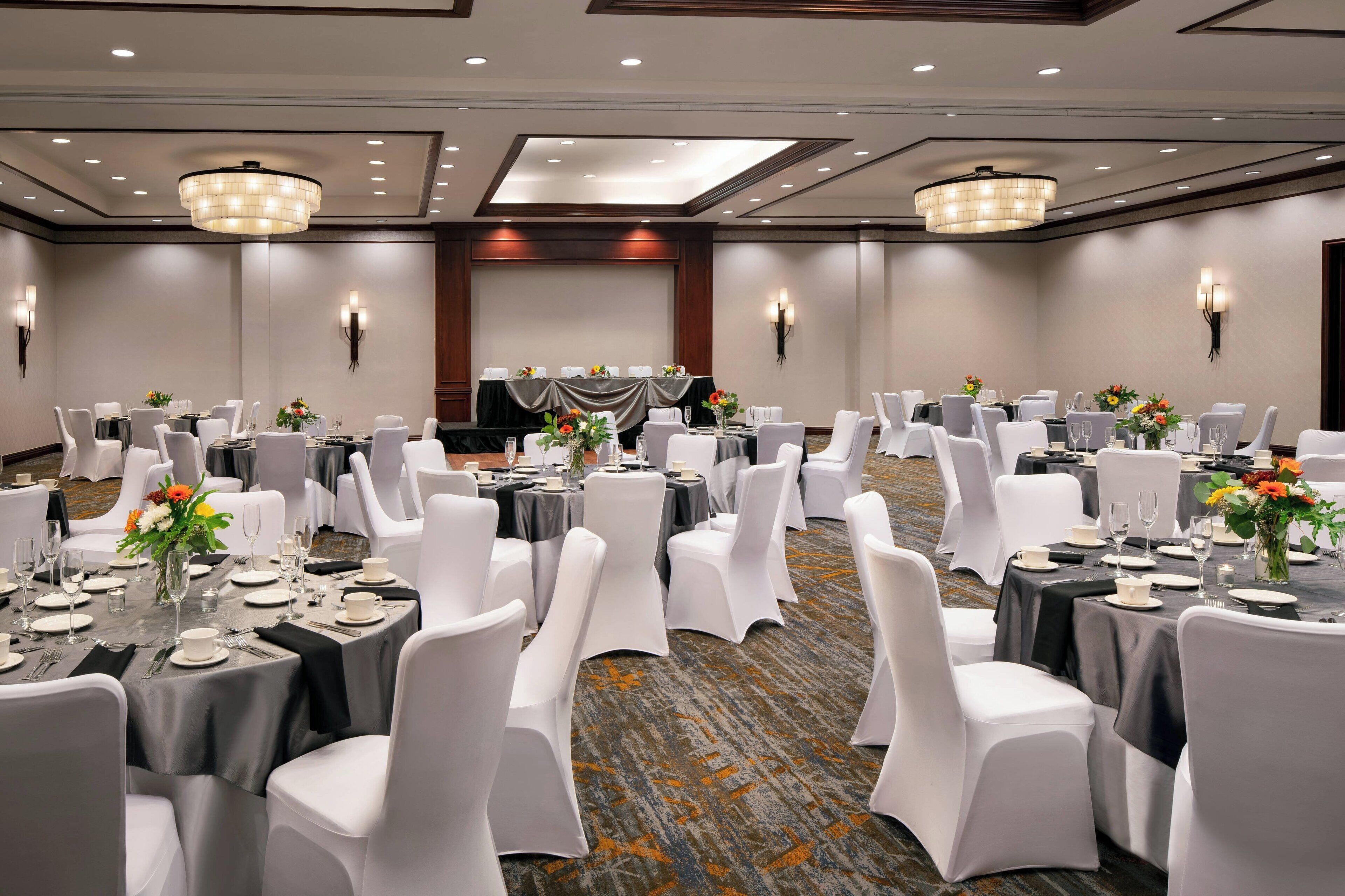 A banquet hall with round tables, white chairs, and chandeliers.