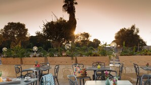 Un área de comedor al aire libre con mesas y sillas, rodeada de plantas y árboles, bajo un cielo despejado.