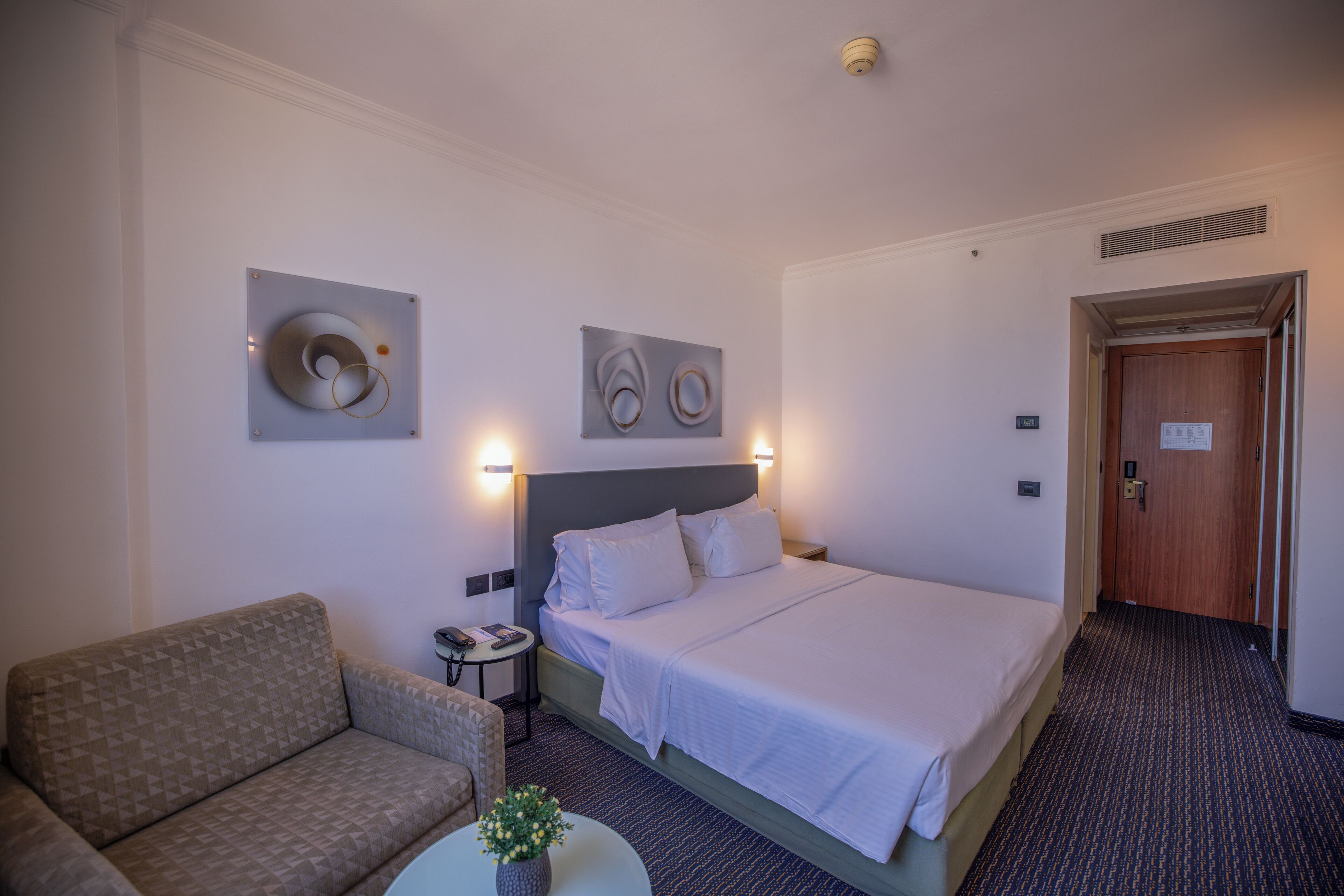 Une chambre d’hôtel comprenant un lit, un canapé, une petite table avec un vase et deux appliques murales.