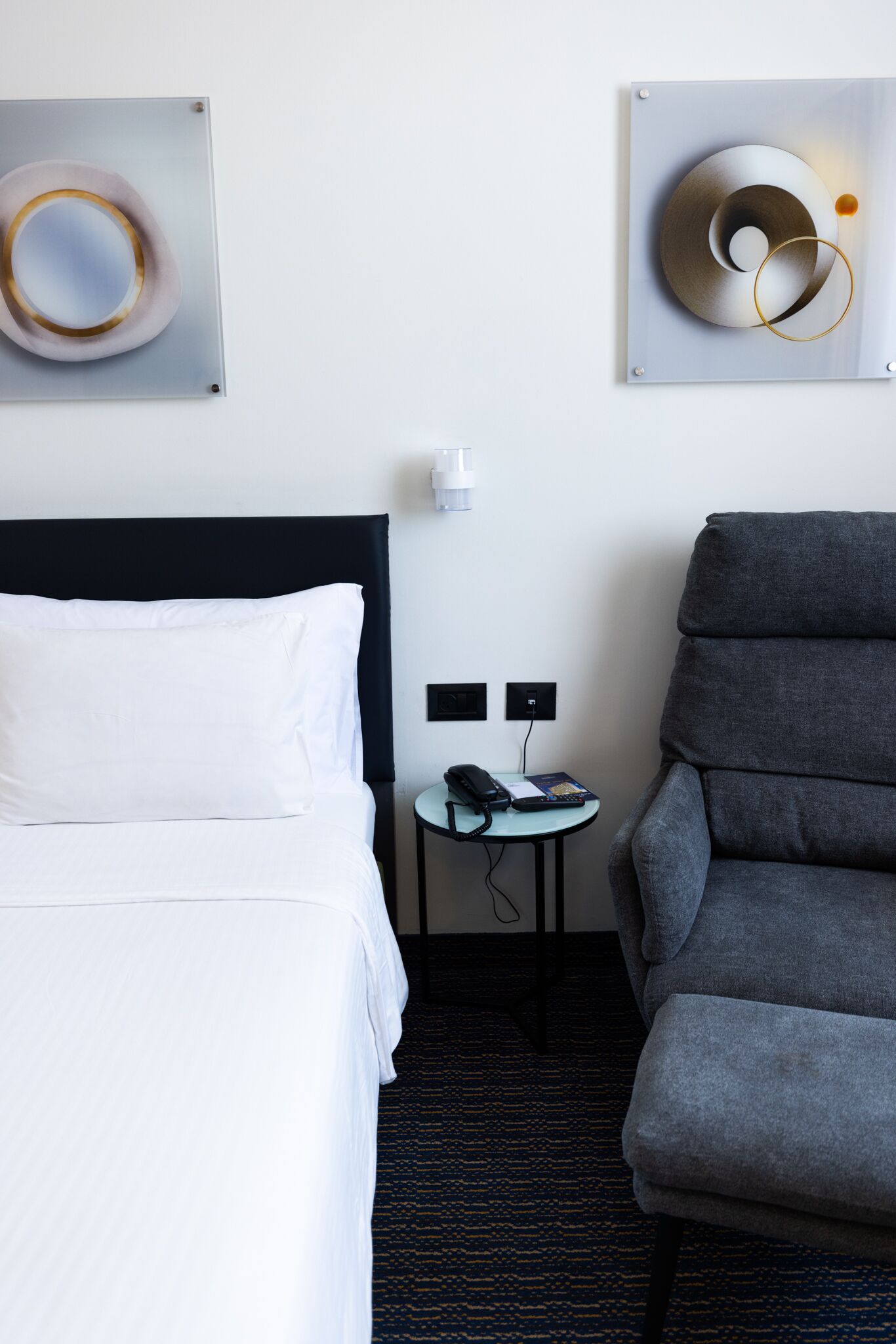 A hotel room with a bed, a grey armchair, a small table with a phone and remote control, and two wall-mounted light switches.