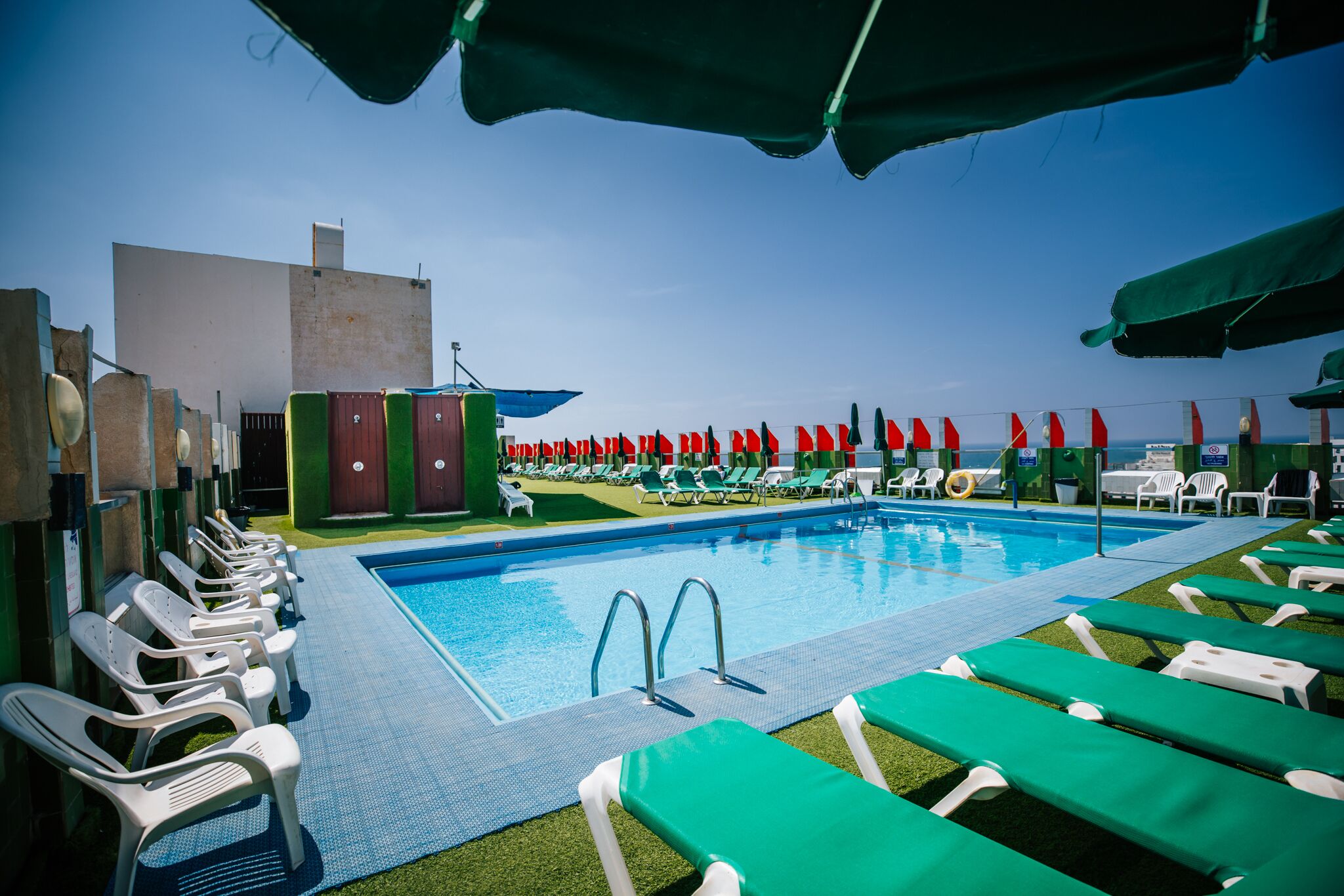 Zona de piscina con sillones de descanso y sombrillas, cielo despejado y un edificio al fondo.