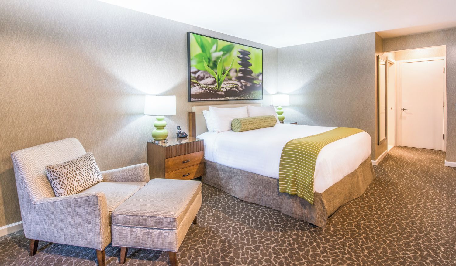 A hotel room with a bed, a chair, a nightstand, and a painting on the wall.