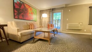 A living room with a sofa, a glass-top coffee table, a chair, a large abstract painting, a window with curtains, and a radiator. - Creekside Inn (Palo Alto)