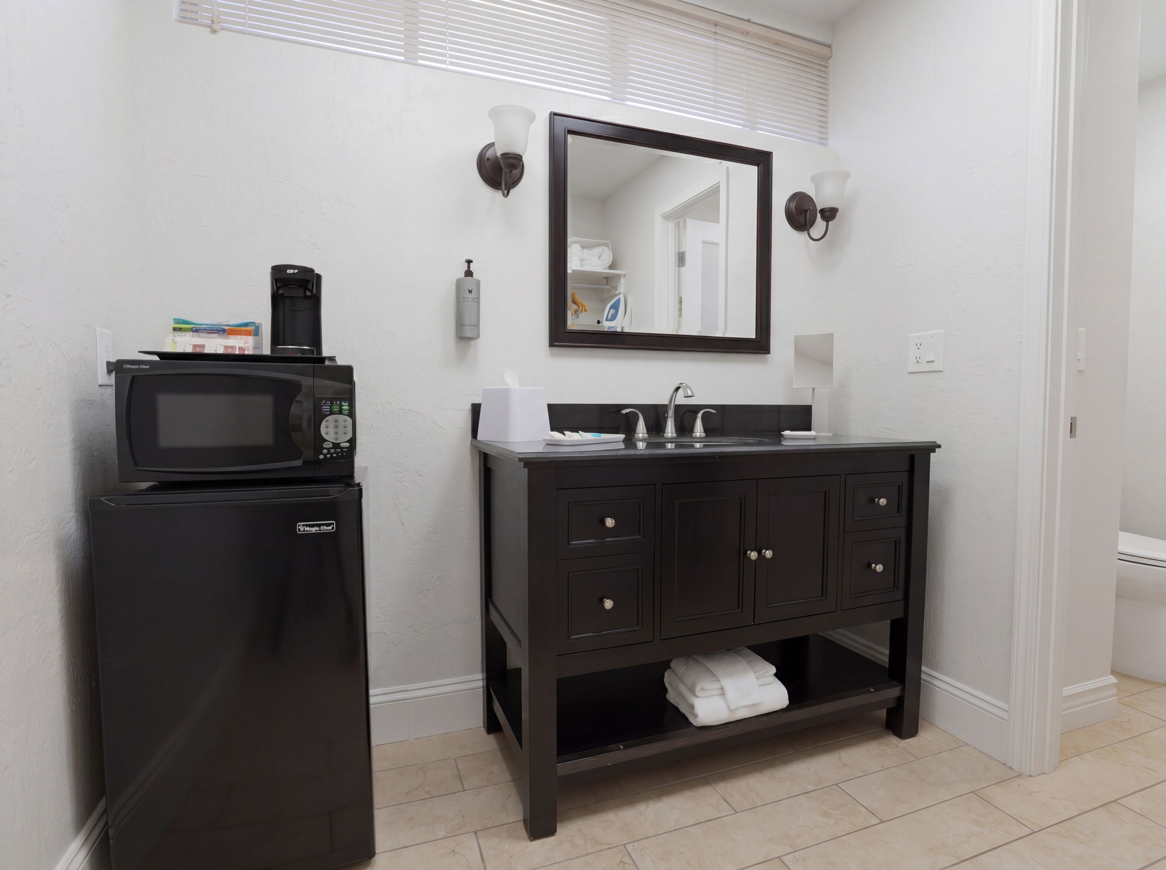 A compact bathroom with a black refrigerator, a sink with a mirror above, and a towel rack below.