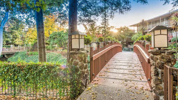 A stone bridge with metal railings leading to a building with a balcony. - Creekside Inn (Palo Alto)