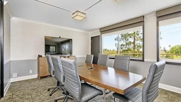 A conference room with a wooden table, six chairs, a television, and large windows with a view of greenery.