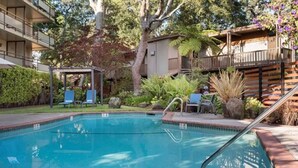 A swimming pool with a wooden deck and lounge chairs, surrounded by greenery and a building with balconies. - Creekside Inn (Palo Alto)