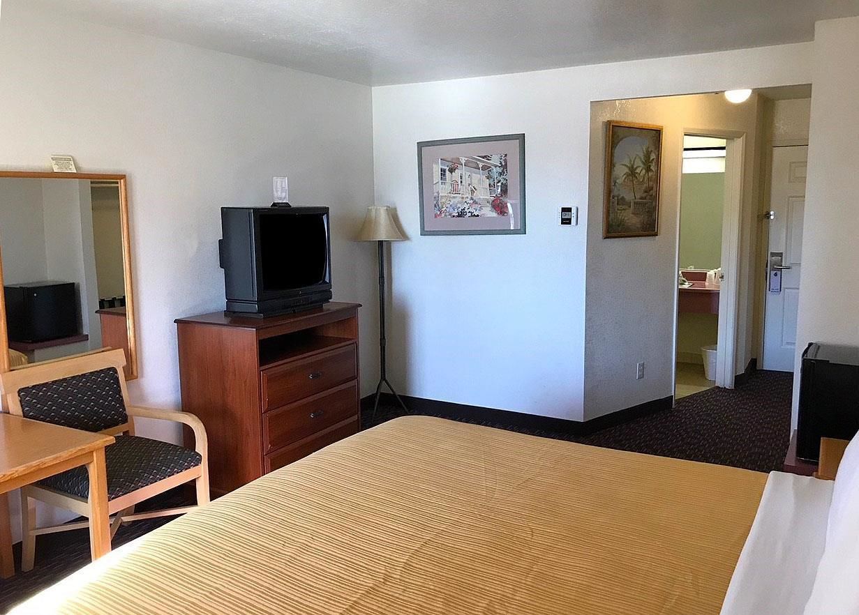 A hotel room with a bed, a desk with a chair, a television on a wooden cabinet, a lamp, and a painting on the wall.