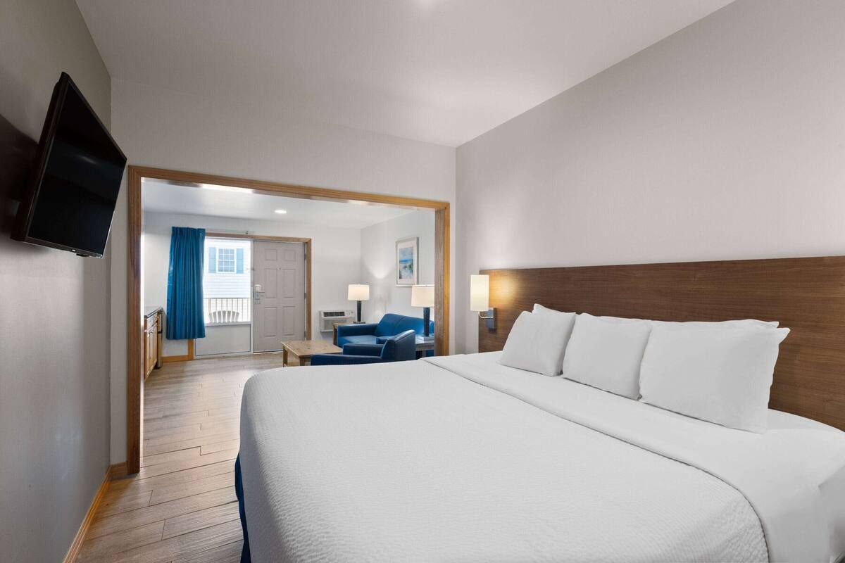 A hotel room with a large bed, a flat-screen TV mounted on the wall, a desk with a chair, and a view of the balcony through an open door.