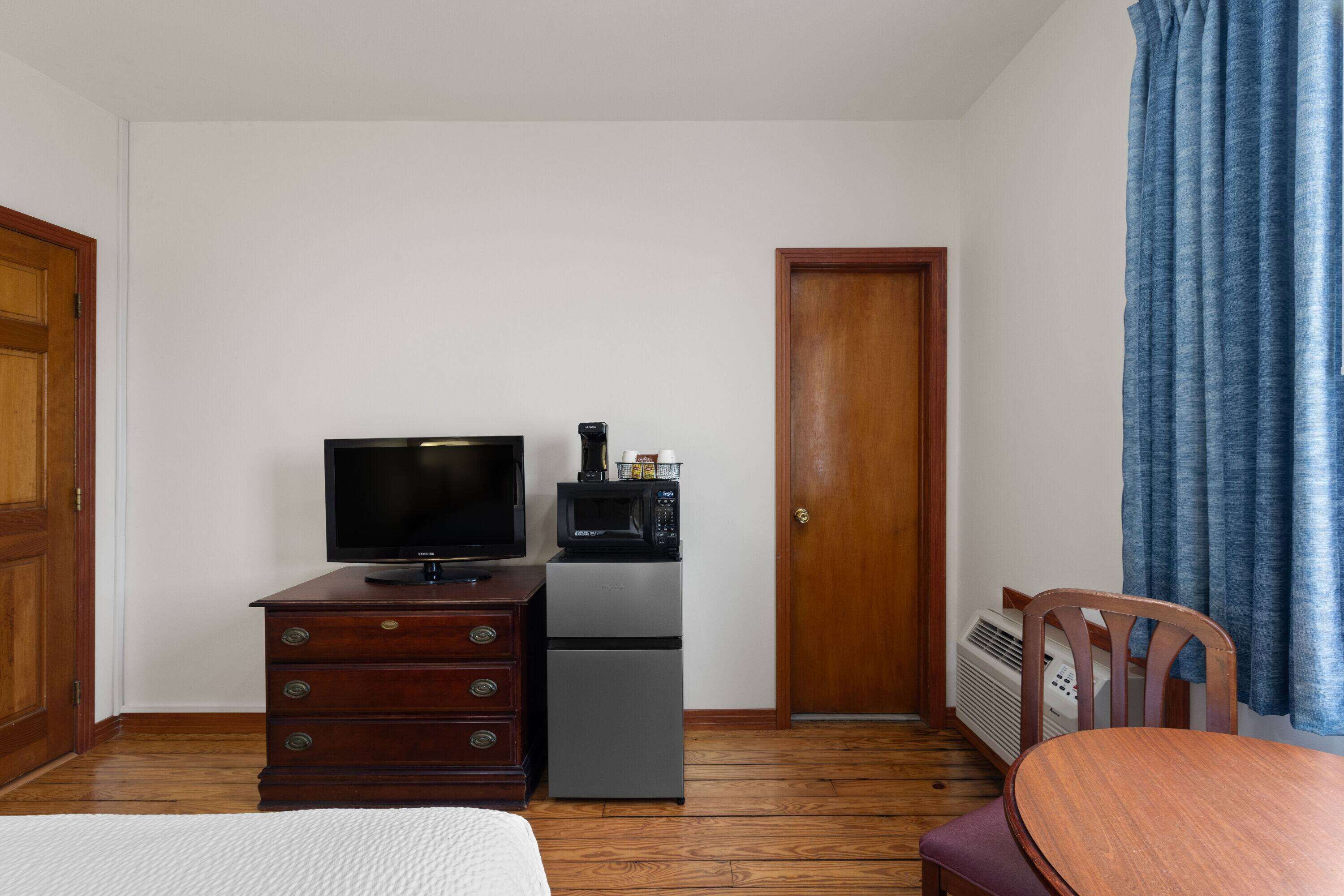 A hotel room with a wooden dresser, a television, a small refrigerator, a wooden chair, and a door.