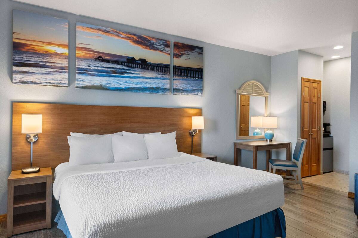 Habitación de hotel con una cama grande, dos lámparas de noche, un escritorio con una silla, un armario de madera y una pared con tres cuadros de paisajes al atardecer.