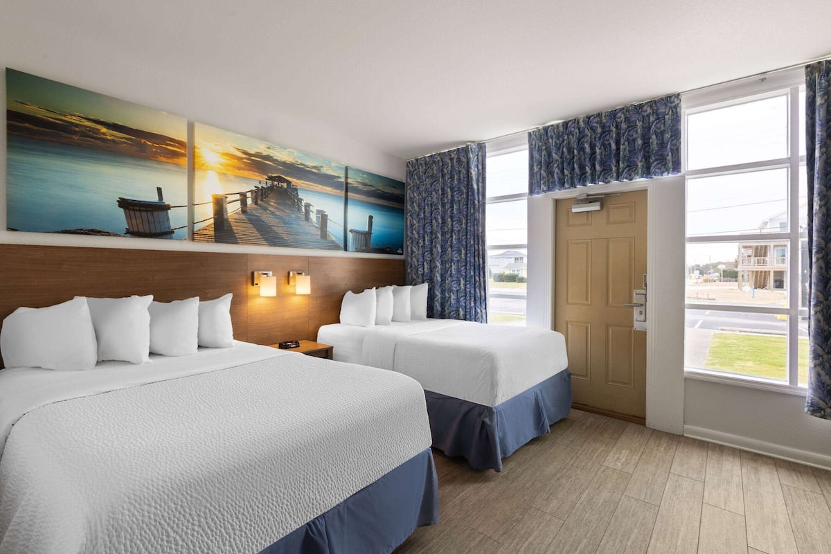 Habitación de hotel con dos camas, un cuadro grande de un atardecer, cabecera de madera y un amplio ventanal con cortinas.