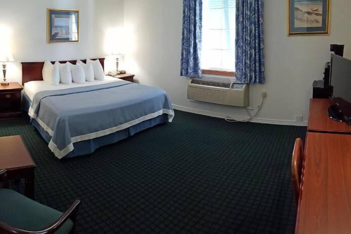 Habitación de hotel con una cama, dos mesitas de noche con lámparas, un escritorio con computadora, una silla, una ventana con cortinas y un aire acondicionado instalado en la pared.