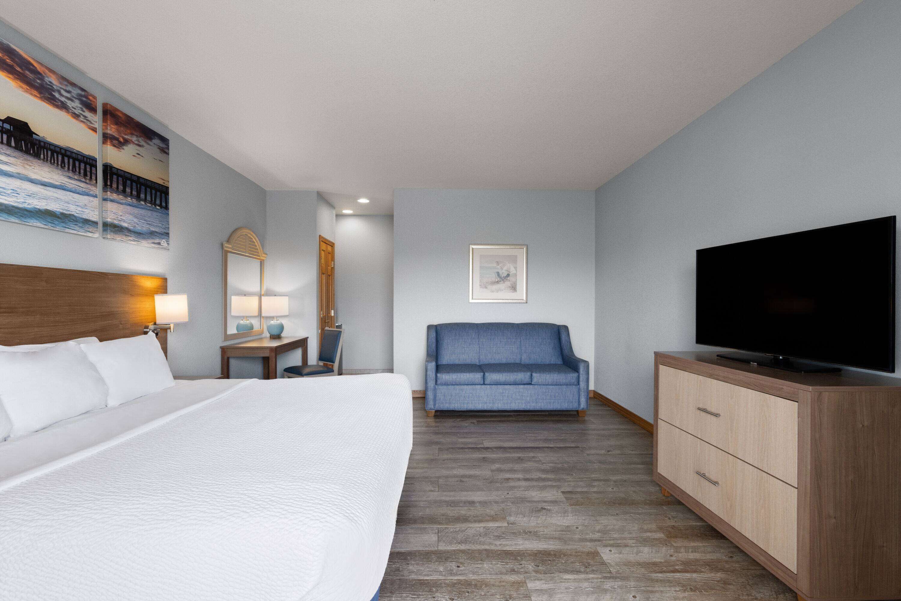 Habitación de hotel con una cama grande, un sofá azul, un aparador de madera, un televisor de pantalla plana y obras de arte en las paredes.