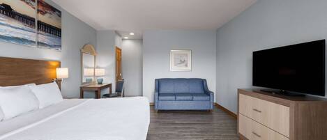 Habitación de hotel con una cama grande, un sofá azul, un aparador de madera, un televisor de pantalla plana y obras de arte en las paredes.
