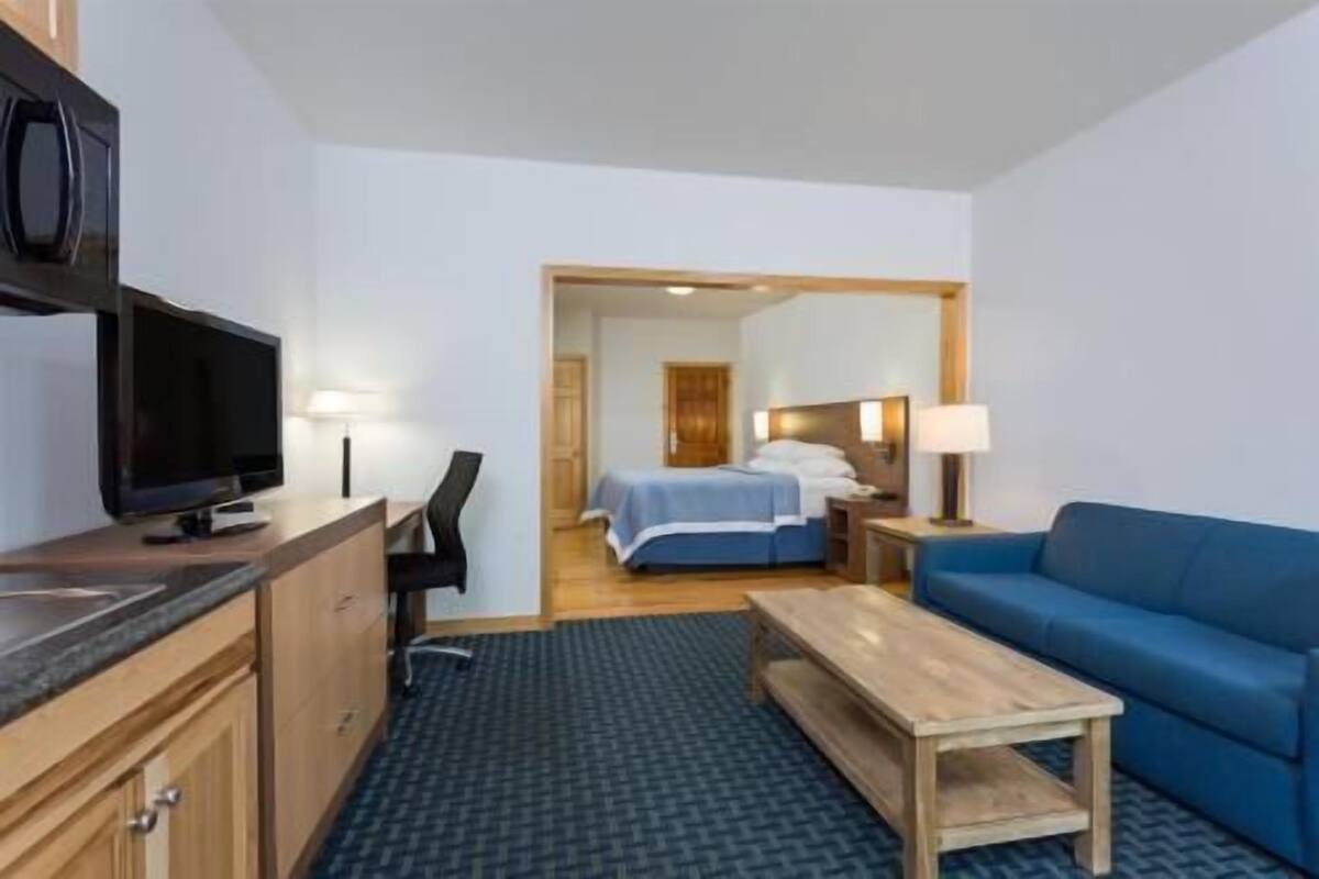 Una habitación de hotel con una cama, un sofá azul, una mesa de centro de madera, un escritorio con una computadora y un televisor.