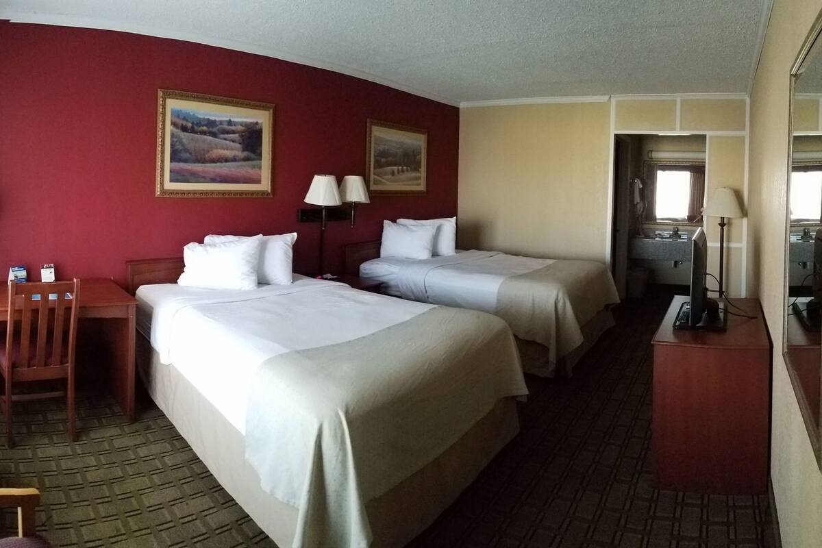 A hotel room with two beds, a desk, a chair, two lamps, and a bathroom visible through an open door.