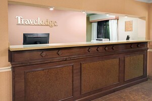 Un mostrador de recepción con un cartel de 'Travelodge', una computadora Lenovo y un tablón de anuncios con varios avisos.