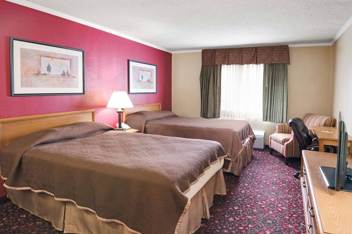 A hotel room with two beds, a desk, a chair, a lamp, and two framed pictures on the wall.