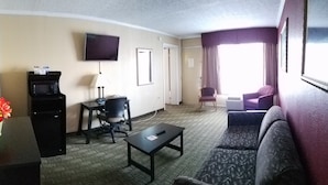 A hotel room with a flat-screen TV, a desk with a lamp, a chair, a sofa, and a large window with curtains. - Travelodge by Wyndham Beckley (Beckley)