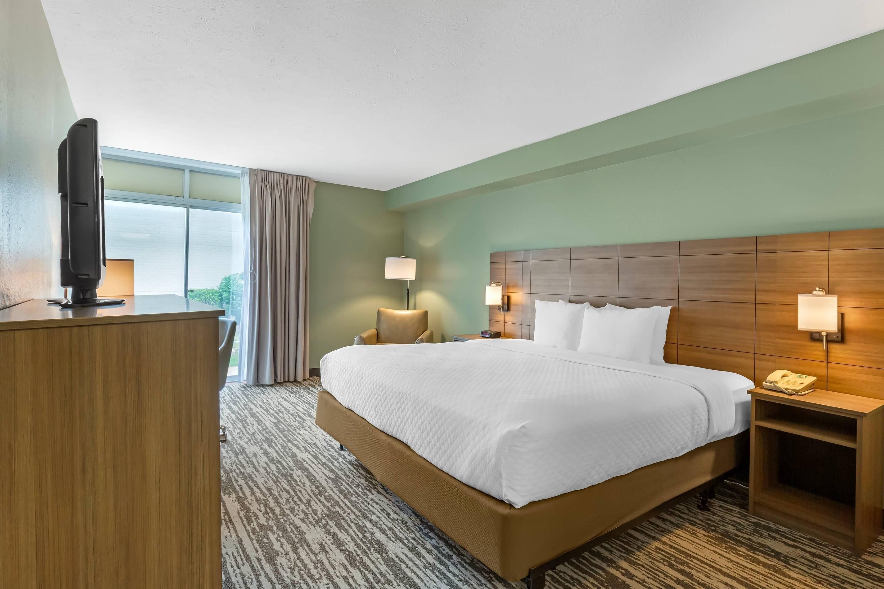 A hotel room with a large bed, a desk, two bedside lamps, a telephone, and a window with curtains.
