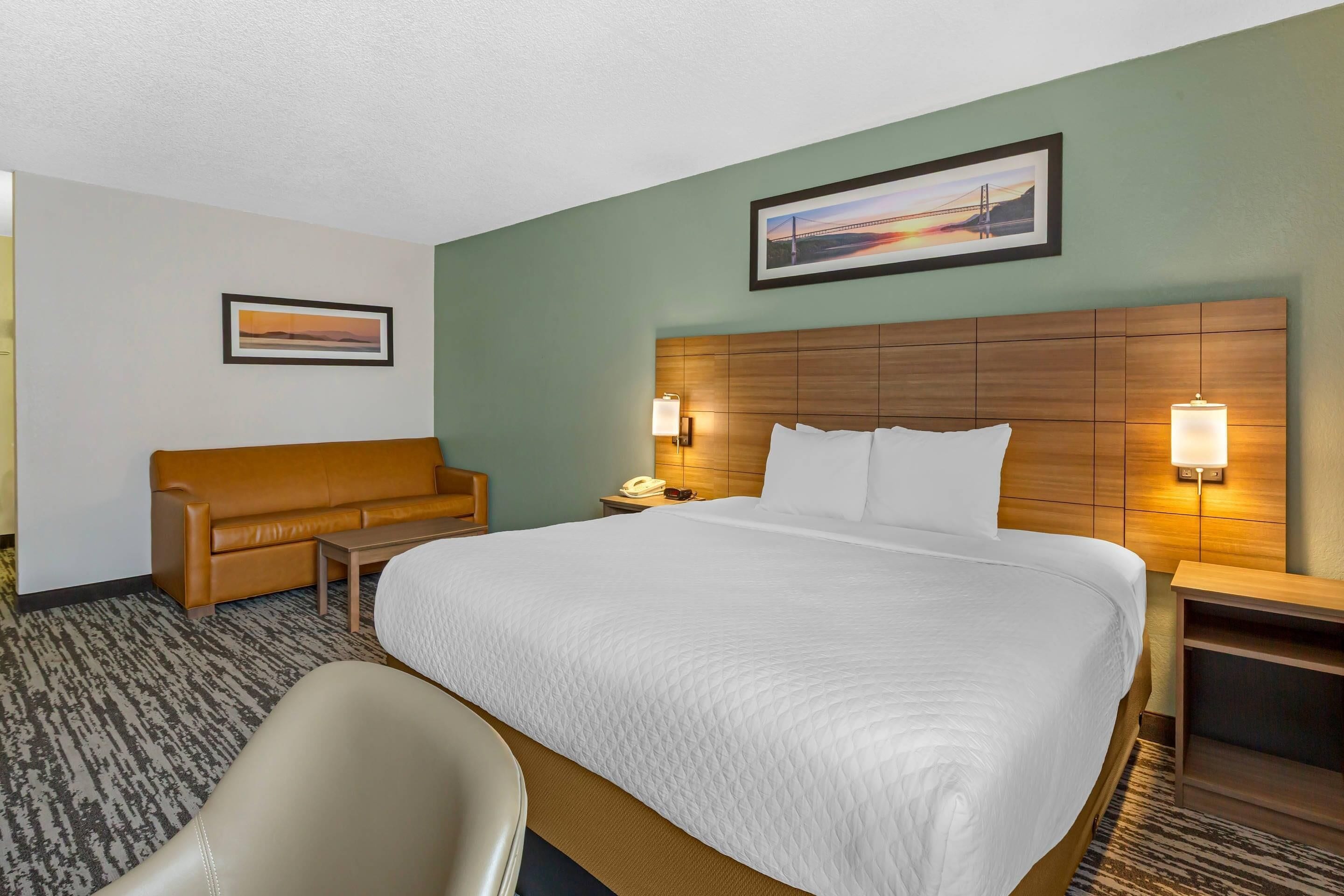 A hotel room with a large bed, a wooden headboard, two bedside lamps, a brown leather sofa, a small table, and a framed picture on the wall.