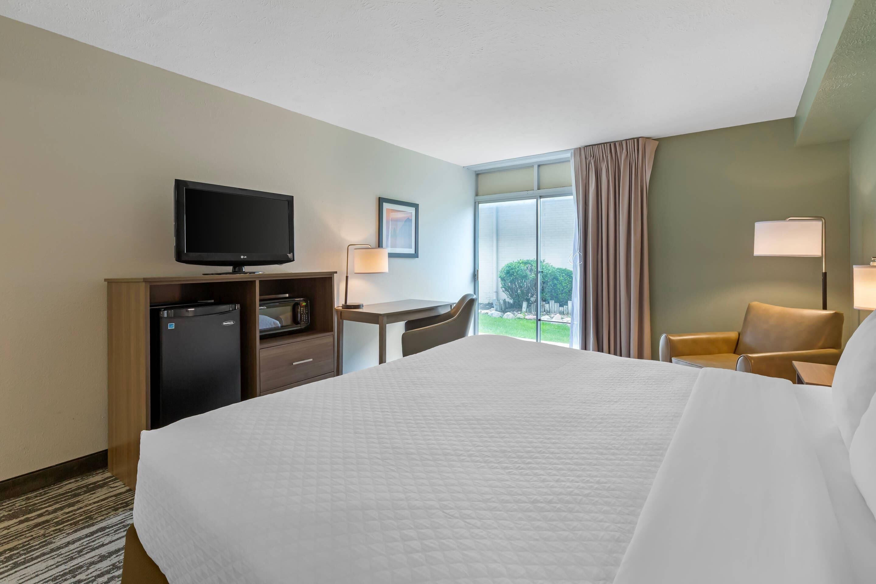 A hotel room with a bed, a desk, a television, a refrigerator, a chair, and a window with curtains.