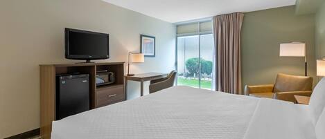 A hotel room with a bed, a desk, a television, a refrigerator, a chair, and a window with curtains.