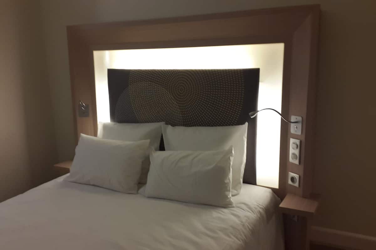 A neatly made bed with white linens and pillows, a headboard with a textured design, and a bedside table with a lamp and a power outlet.
