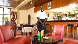 A bar area with a bartender, red leather chairs, a round table with drinks, and a statue. - Hôtel La Tchadienne (Ex Novotel) (N'Djamena)