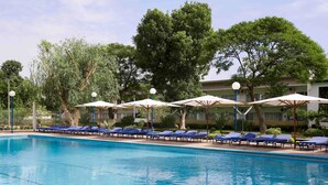A swimming pool with blue lounge chairs and umbrellas, surrounded by trees and a building in the background. - Hôtel La Tchadienne (Ex Novotel) (N'Djamena)