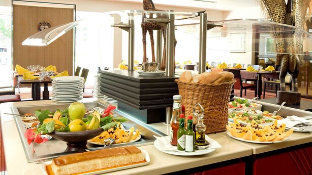 A buffet with various food items, including fruits, bread, and a cake, with a giraffe and elephant sculpture in the background. - Hôtel La Tchadienne (Ex Novotel) (N'Djamena)