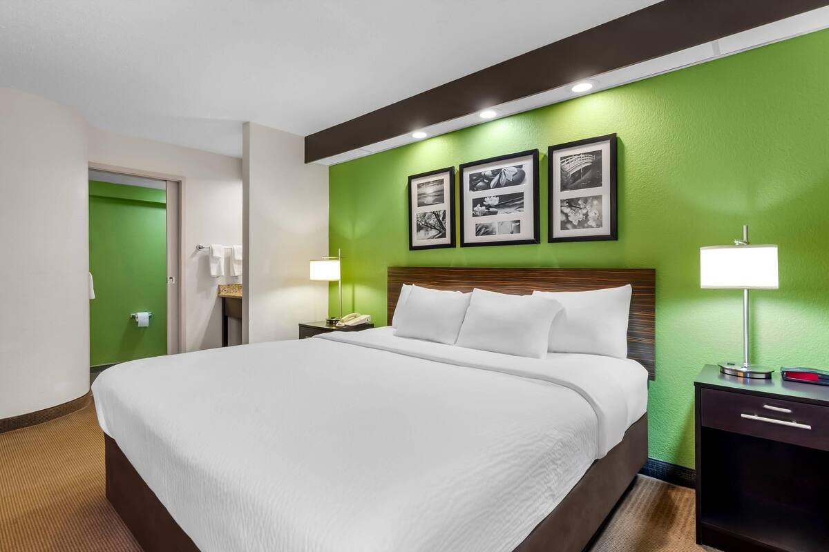 A hotel room with a large bed, a nightstand with a lamp, a green accent wall, and framed pictures above the bed.