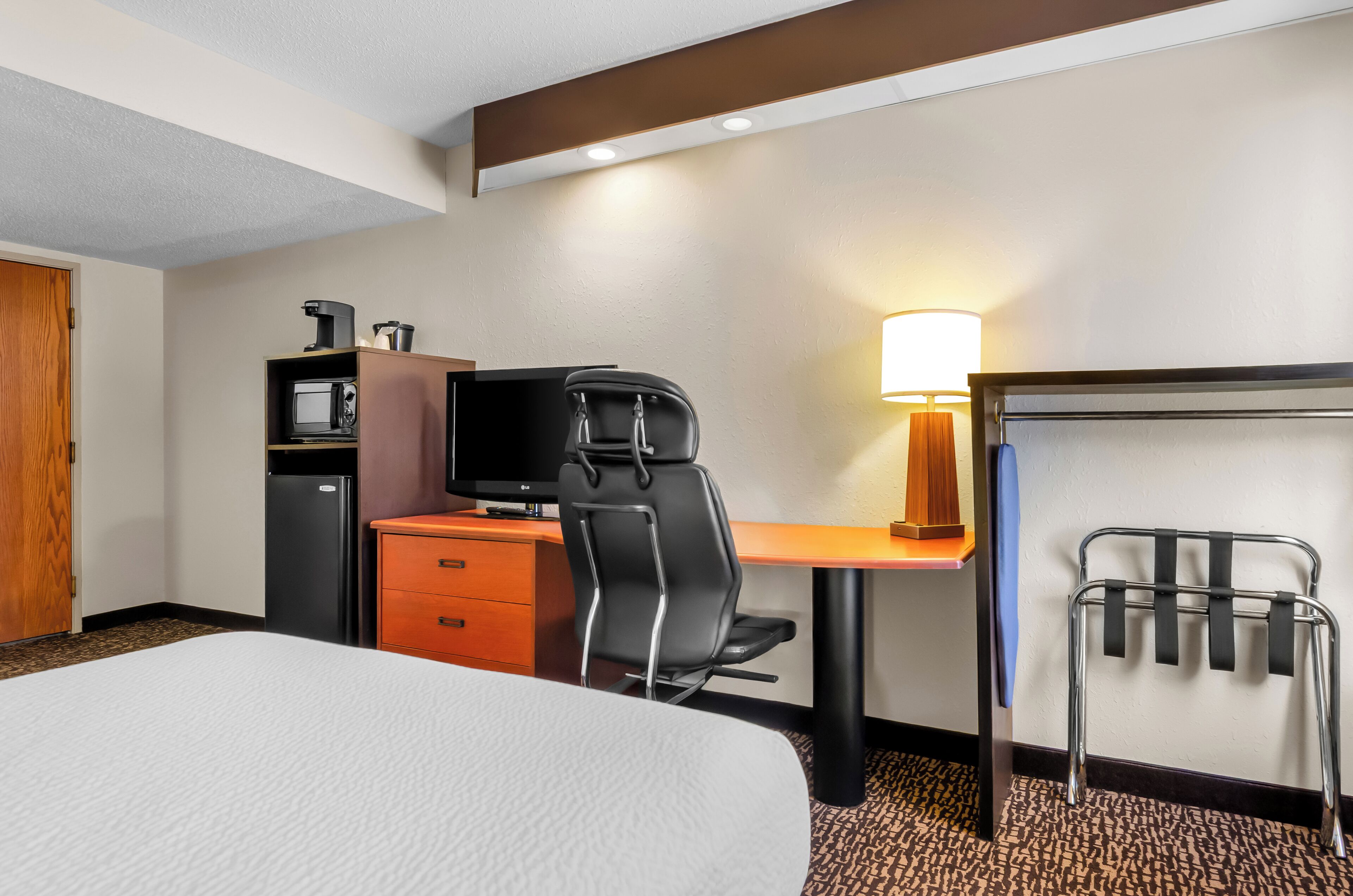 Habitación de hotel con cama, escritorio con computadora y lámpara, silla, refrigerador pequeño y microondas.