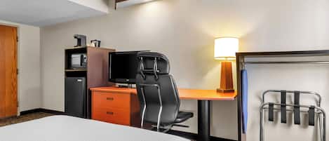 Habitación de hotel con cama, escritorio con computadora y lámpara, silla, refrigerador pequeño y microondas.