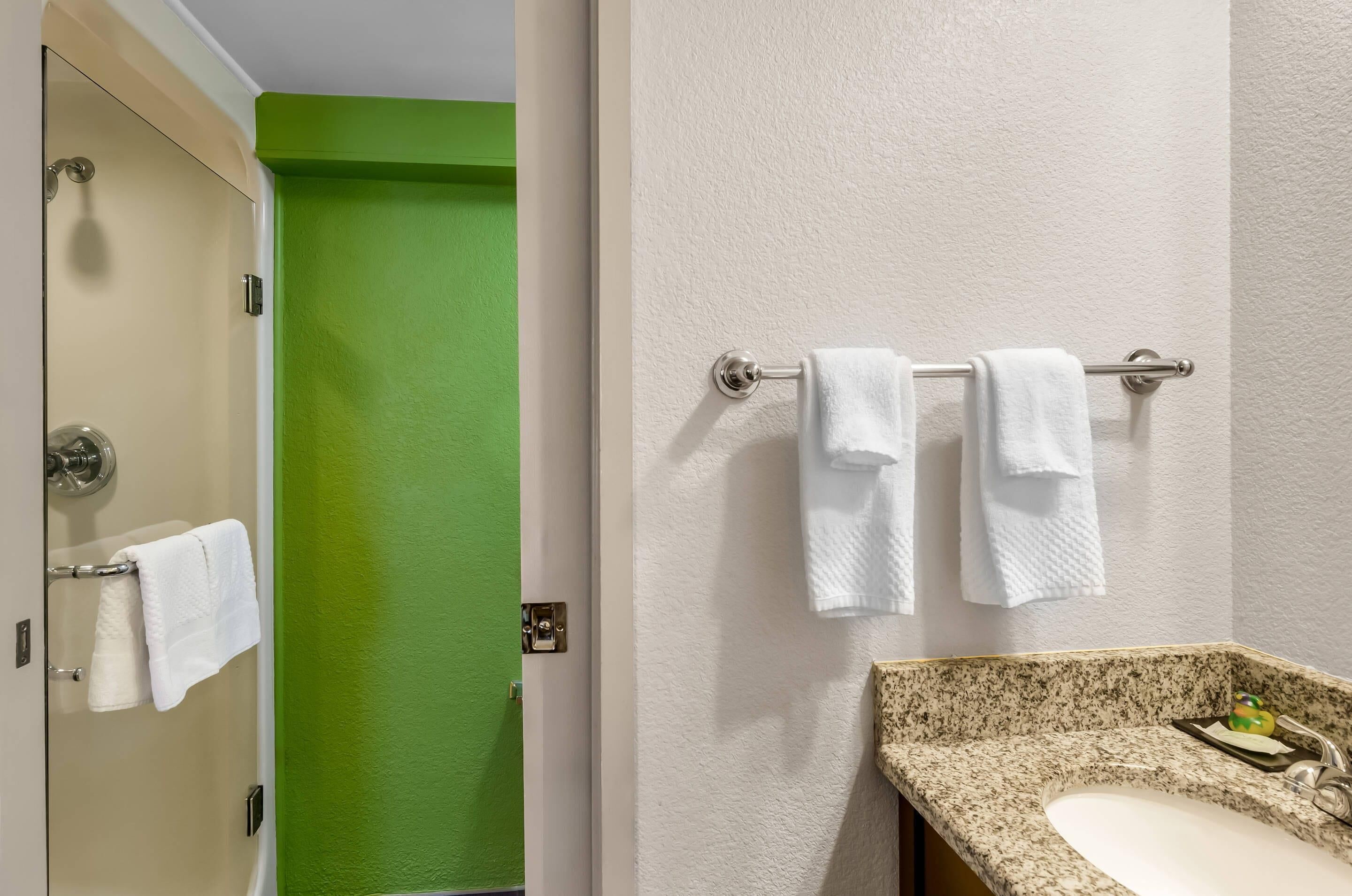 Une salle de bain avec une porte verte, un lavabo blanc doté d’un plan de travail en granit, et deux serviettes blanches suspendues à un porte-serviettes.