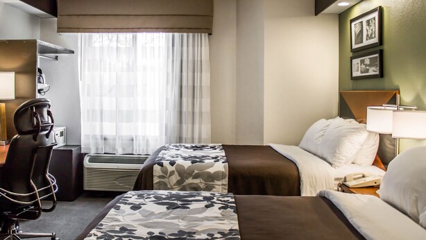 Une chambre d’hôtel avec deux lits, un bureau, une chaise, un appareil de climatisation et une fenêtre avec des rideaux légers.