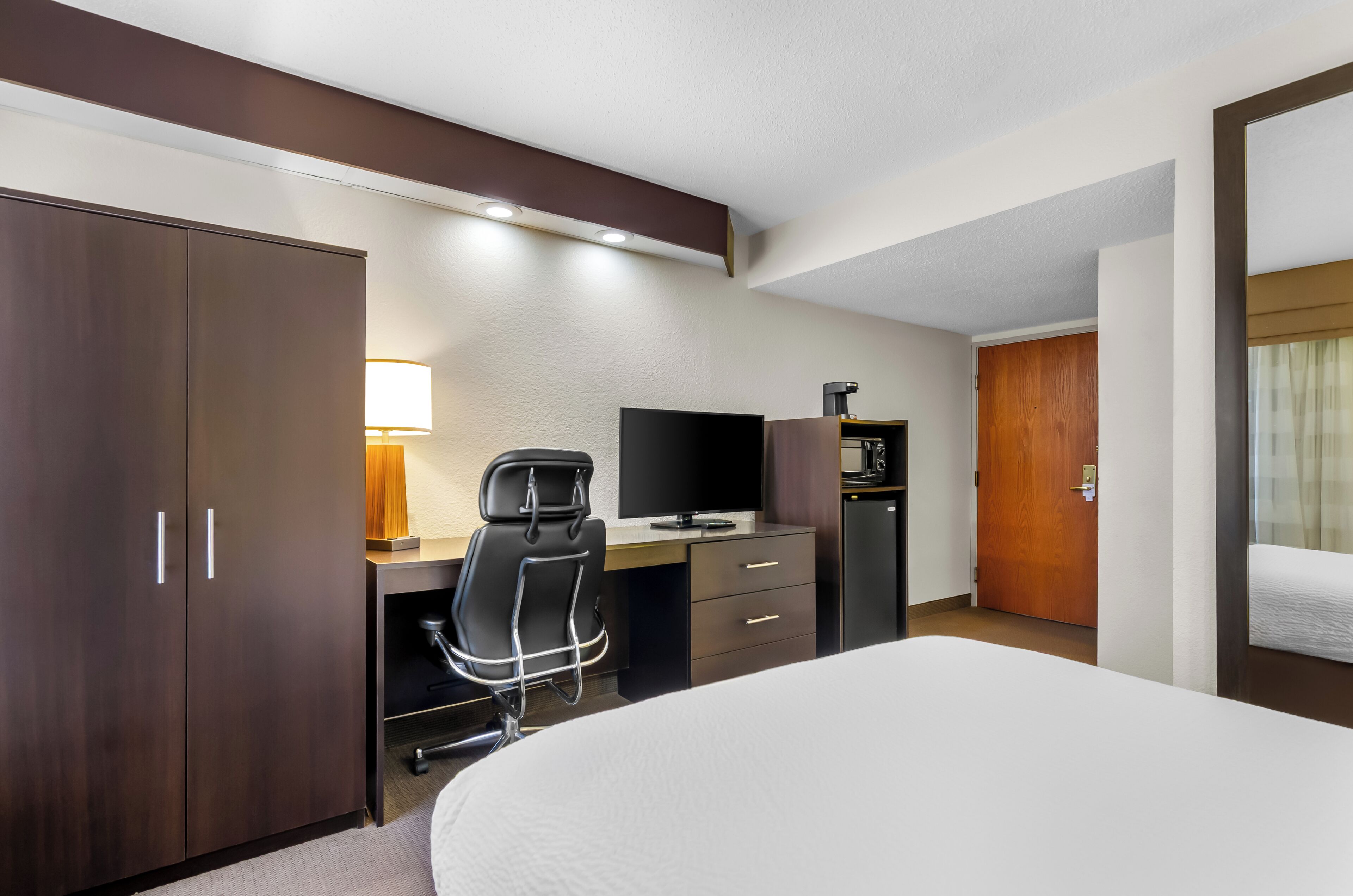 A hotel room with a bed, a desk with a chair, a TV, a minibar, and a wardrobe.