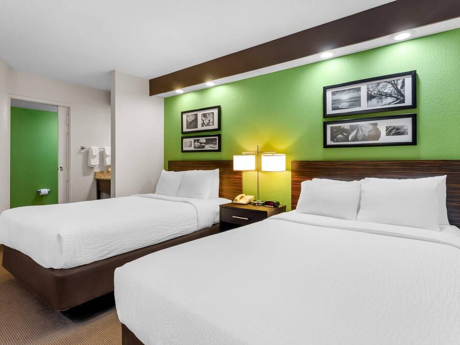 A hotel room with two beds, a green accent wall, framed pictures, a nightstand with a lamp, and a phone.