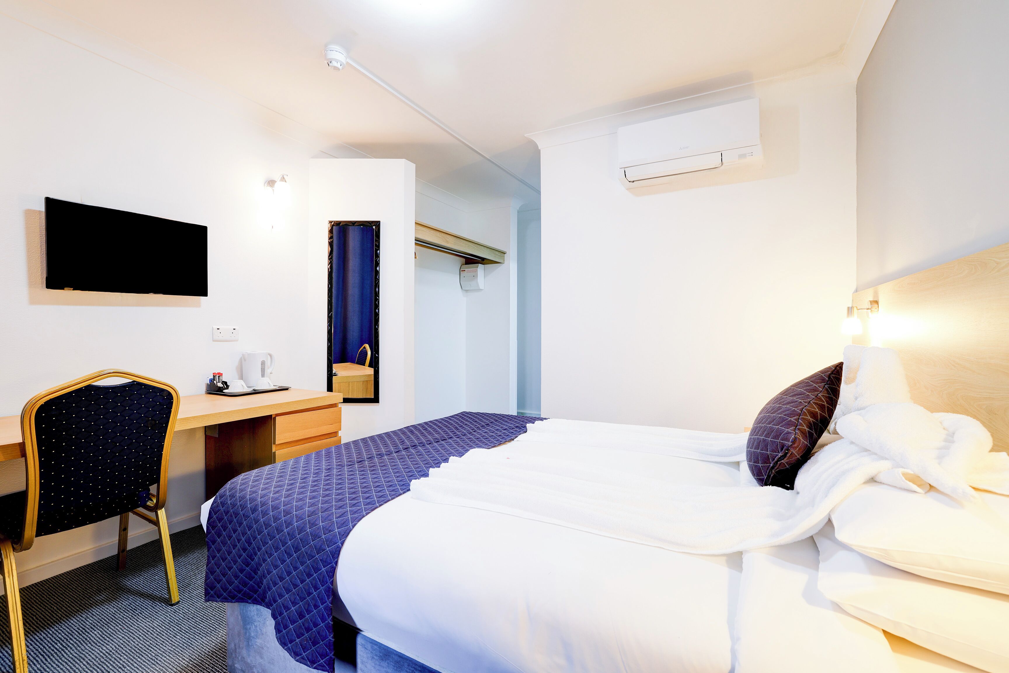 A hotel room with a bed, a desk, a chair, a flat-screen TV, and an air conditioning unit.