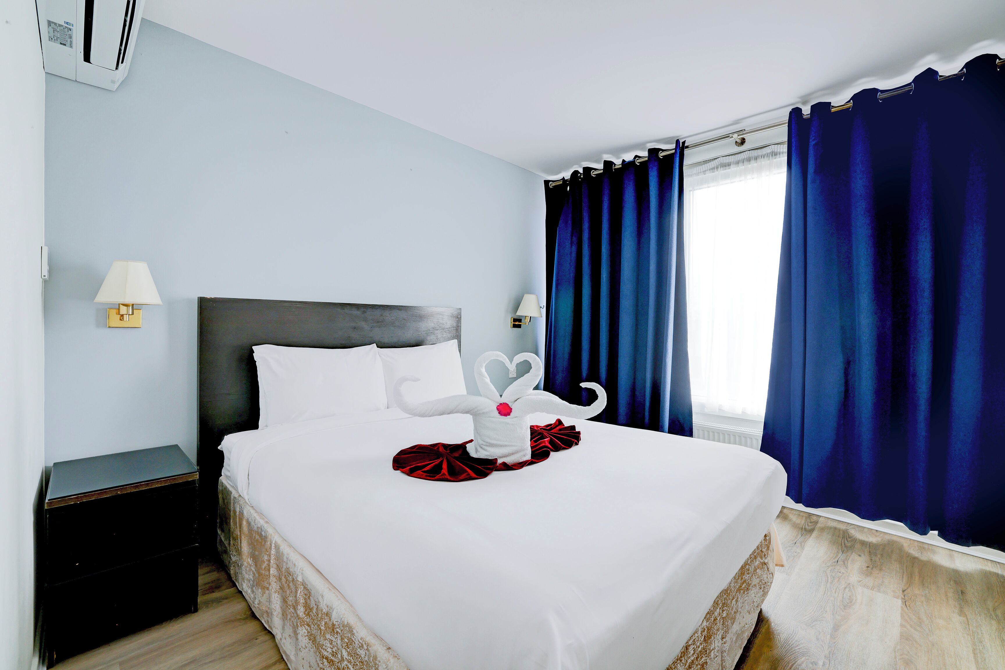 Una cama bien hecha con sábanas blancas y una decoración de toallas. Hay una mesita de noche pequeña con una lámpara y un buró de color oscuro. La habitación tiene paredes azul claro, un cabecero oscuro y una ventana con cortinas azules.