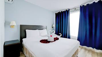 A neatly made bed with white linens and a decorative towel arrangement. There is a small bedside table with a lamp and a dark-colored nightstand. The room has light blue walls, a dark headboard, and a window with blue curtains.