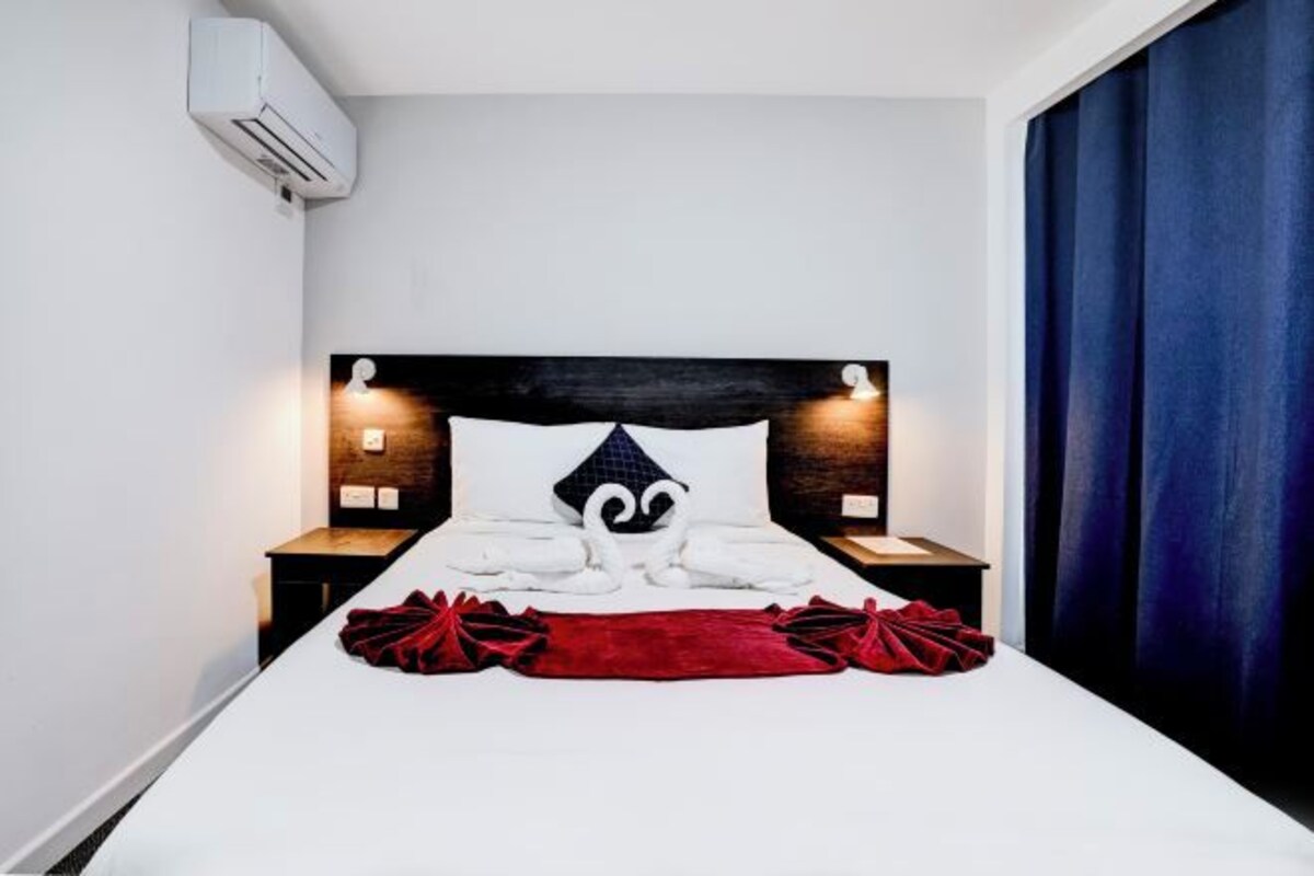 A modern hotel room with a large bed, a headboard with integrated lighting, a red throw blanket, and a blue curtain.