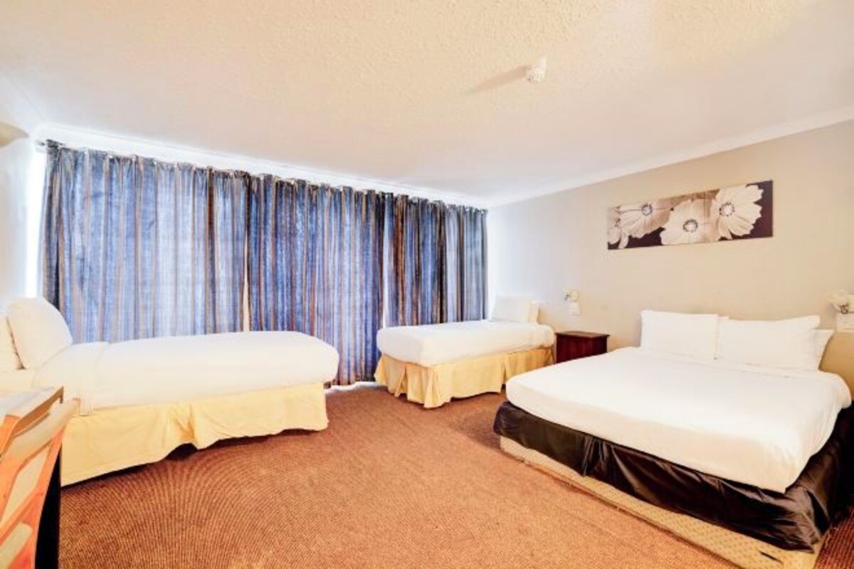 A hotel room with two single beds, a large bed, a window with curtains, a painting on the wall, and a carpeted floor.
