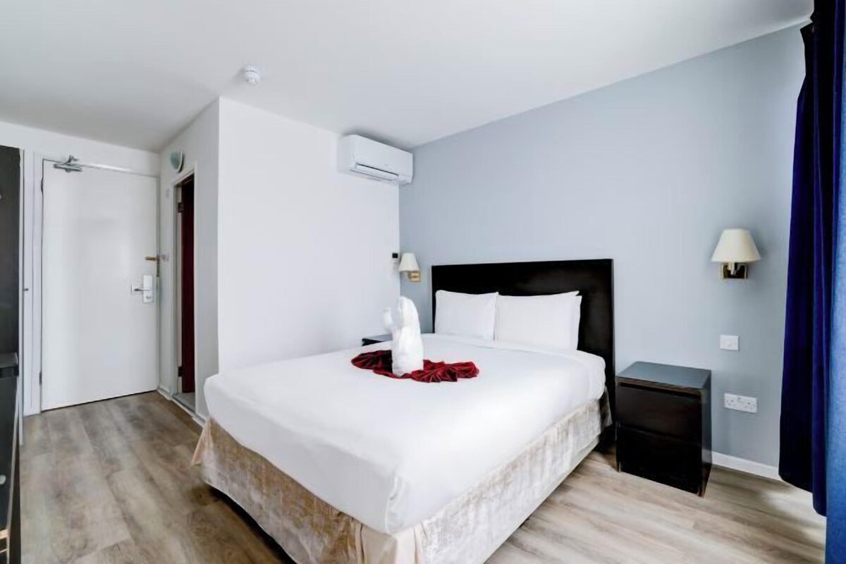 A hotel room with a large bed, a bedside table, an air conditioning unit, and a lamp.