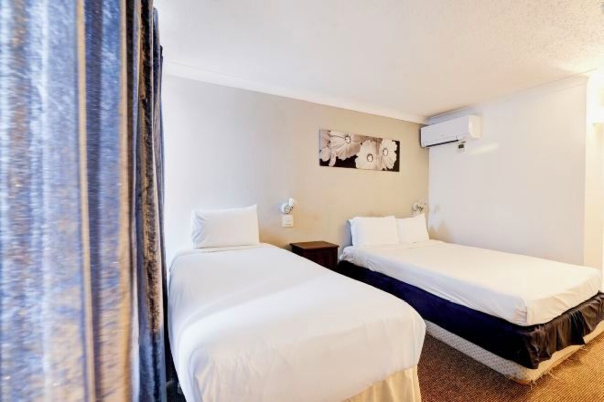 A hotel room with two single beds, a small bedside table, a wall-mounted air conditioner, and a picture of flowers on the wall.