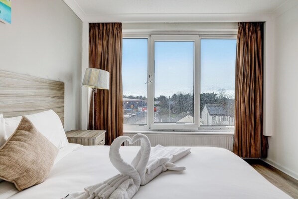 A hotel room with a bed, a heart-shaped towel, a lamp, and a window with curtains.