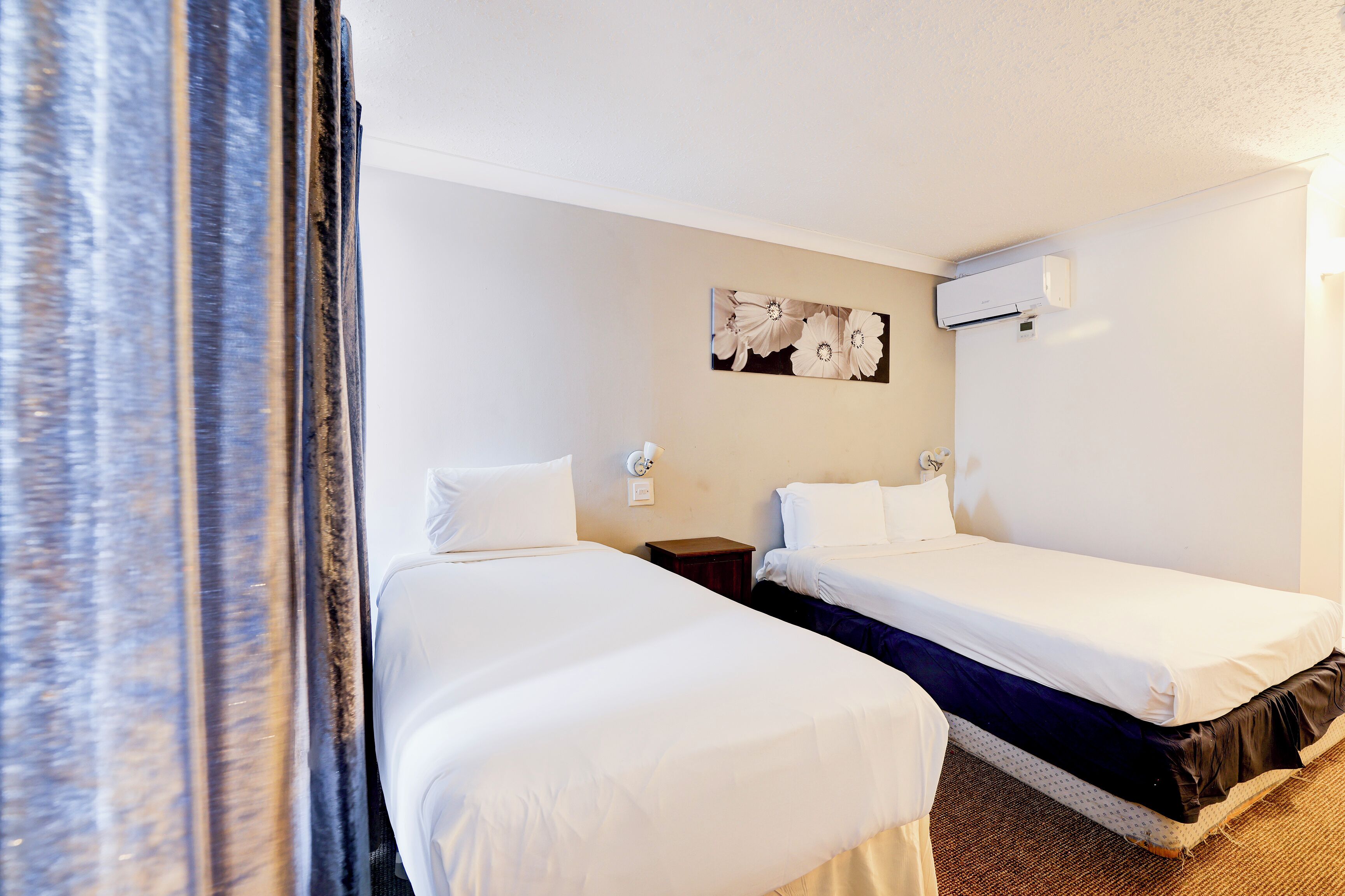 Ein Hotelzimmer mit zwei Einzelbetten, einem Nachttisch, einer Wand-Klimaanlage und einem Blumengemälde.