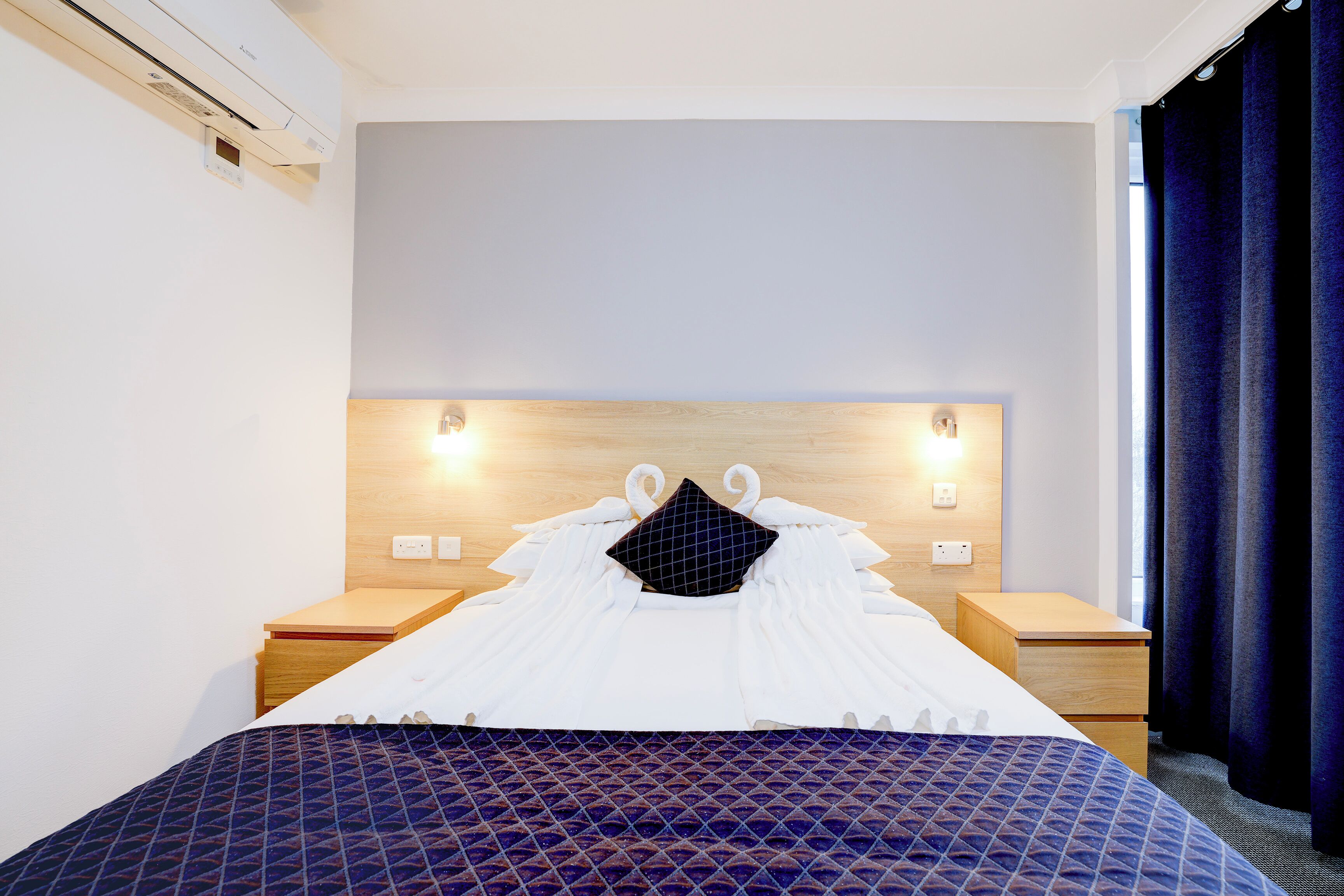 Een hotelkamer met een bed, een houten hoofdeinde, nachtkastjes en een raam met gordijnen.