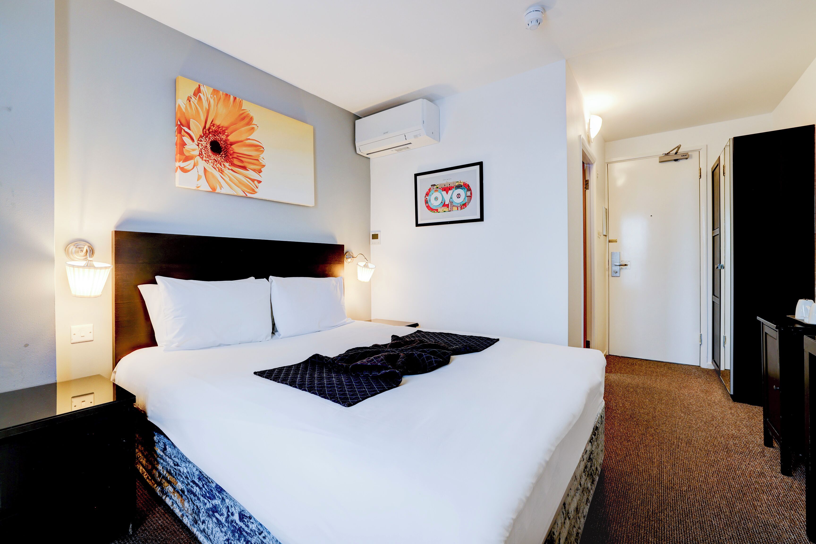 A hotel room with a large bed, a wall-mounted air conditioner, a framed artwork, and a door leading to another room.